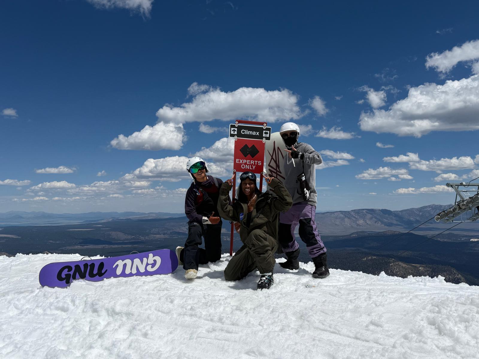 Three snowboarders celebrating at a mountain summit sign on a sunny day