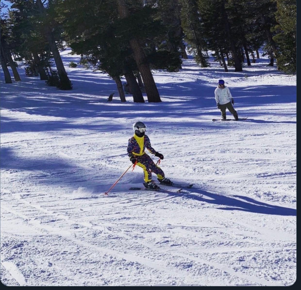 A young skier practicing a controlled turn while an instructor follows behind on a tree-lined slope